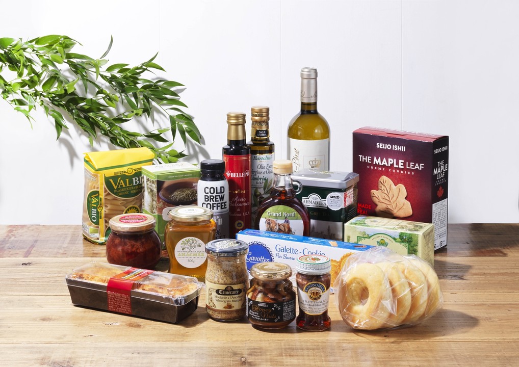 テーブルの上に並べられた、さまざまな輸入食品のセットが写っている。オリーブオイルやワイン、バルサミコ酢、ジャム、クッキー、パン、マスタード、はちみつ、紅茶などの高級食材が整然と並べられている。背景には観葉植物が置かれており、明るくナチュラルな雰囲気を演出している画像。