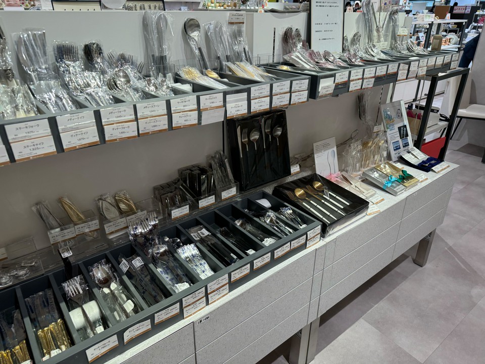 アクセルジャパン カトラリー売り場のアップ。銀色や金色、透明なプラスチックハンドルのフォーク、スプーン、ナイフが整然と並べられている