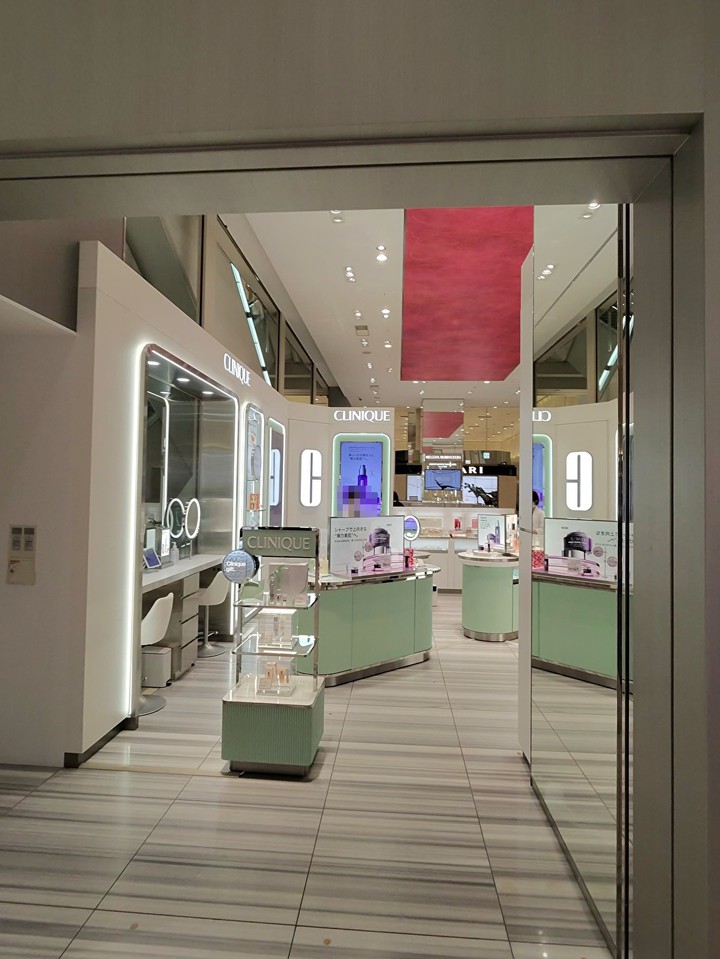 クリニークのエレガントな美容カウンター、中央に配置された商品の展示台と、壁面に取り付けられた鏡とサンプルテスターが特徴。ショップの入り口から見て、天井から吊り下げられた赤いバナーが視界に入る。床は白と灰色のストライプ模様で、モダンなショッピングスペースを演出している。