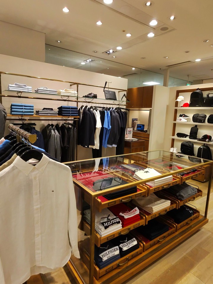TOMMY HILFIGERの店内で、金色のフレームとガラス製ディスプレイテーブルの上に、畳まれたHILFIGERブランドのTシャツが並べられている。背景には、シャツとジャケットが掛けられており、アクセサリーが展示されている棚もある。
