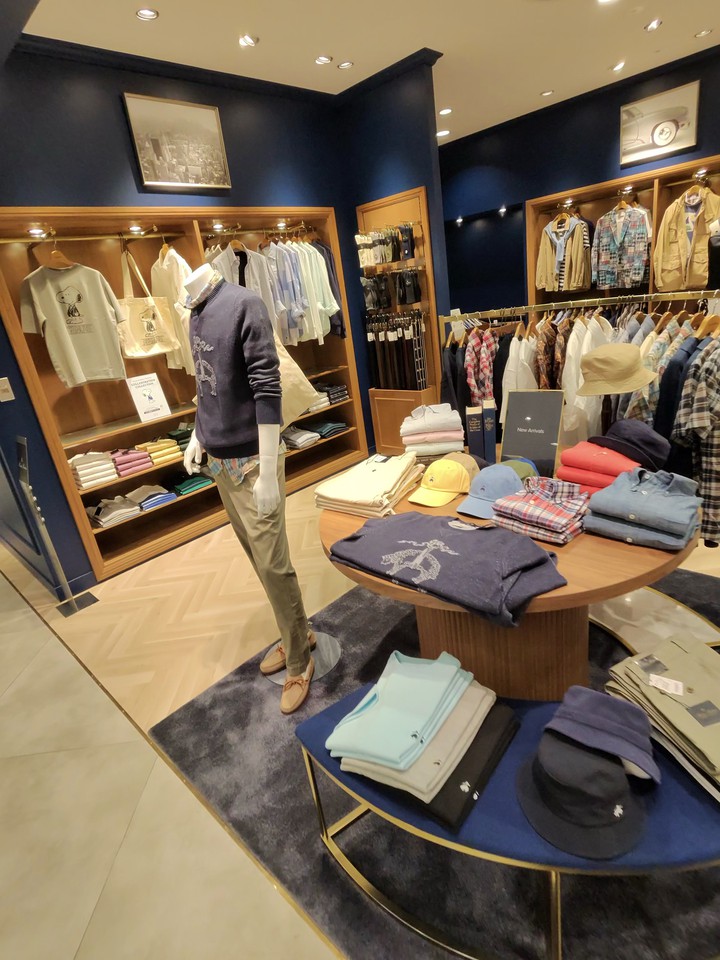 エレガントなブルックスブラザーズの店内で、マネキンがネイビーブルーのセーターとベージュのパンツを着こなしている。背景には白やライトブルーのシャツとニットウェアが丁寧に陳列されており、前面のディスプレイテーブルには折り畳まれた服と帽子が展示されている。