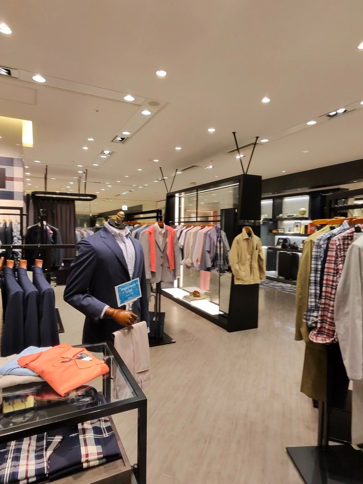 スーツとカジュアルウェアが展示された店内。手前のマネキンはグレースーツにオレンジのセーターを着こなし、手にはブルーの冊子を持っている。背景には多彩なカラーのシャツが並ぶ。