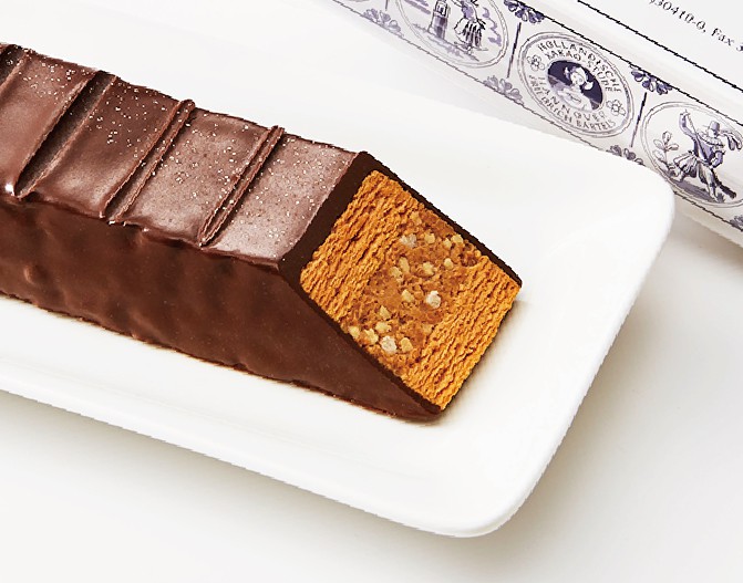 白い皿の上に、チョコレートでコーティングされた長方形のケーキが置かれている。ケーキの断面が見えており、層になった生地の中にサクサクとしたクランチが入っている。上品なチョコレートの光沢が特徴的で、高級感のあるスイーツの画像。