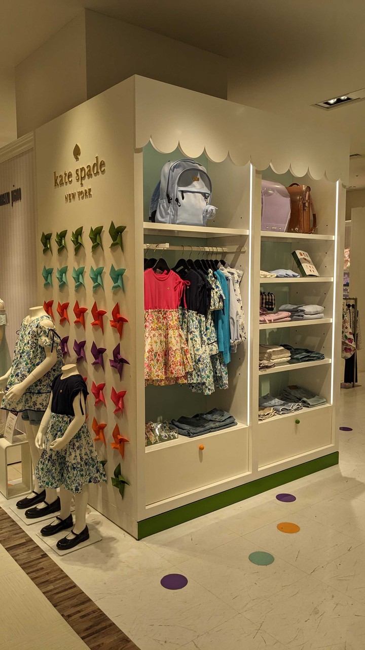 kate spade new york kidsの売り場で、カラフルなピンホイールをバックにした子供服のマネキンと、フローラルプリントのドレスやポロシャツが並んでいる様子です。
