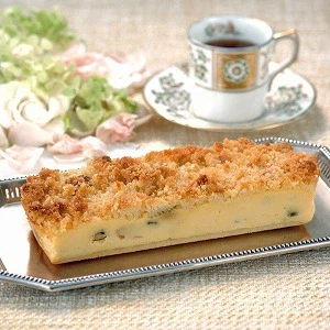 銀のトレーに載せられたクランブルがのったケーキがテーブルに置かれており、その奥にはエレガントなデザインのカップとソーサーに入った紅茶がある。背景には淡い色の花が飾られ、上品なティータイムの雰囲気を演出している画像。