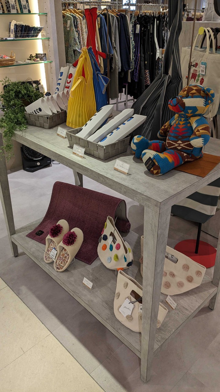 新宿高島屋 エプロン・スリッパ売り場。パッチワークのテディベア、カラフルなストライプのバッグ、そして手作り感のあるスリッパが木製テーブルにディスプレイされている