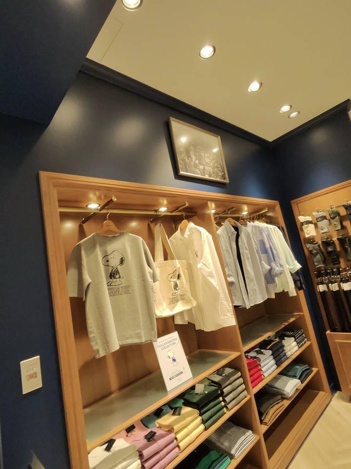 ブルックスブラザーズの店内で、木製のシェルフにはキャラクターがプリントされたグレーとホワイトのTシャツが掛かっている。下の棚にはカラフルなポロシャツが整然と並べられており、店内は落ち着いた照明で照らされている。