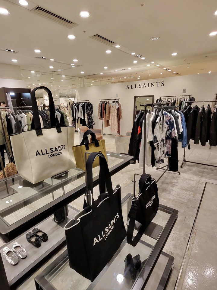 ALLSAINTSのブランドが印字された様々なサイズの黒とベージュのトートバッグがガラスのディスプレイテーブルに展示されている。背景にはALLSAINTSのロゴと衣服が掛かるラックがある。