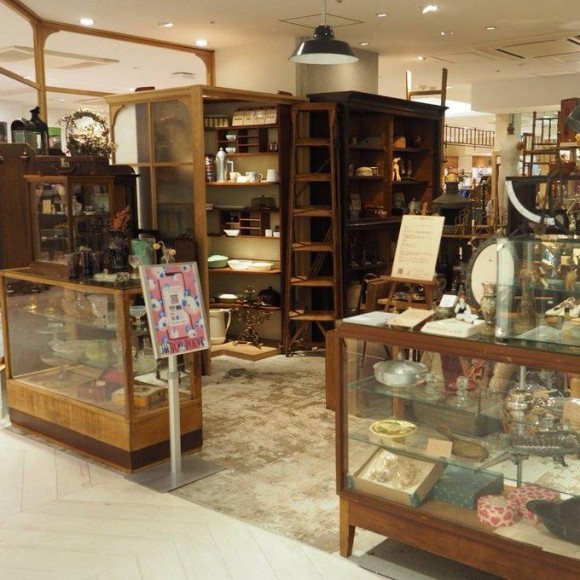 アンティークス・エデュコの展示エリア。ガラスのショーケースや木製の棚に、古風な食器や雑貨が多数展示されている