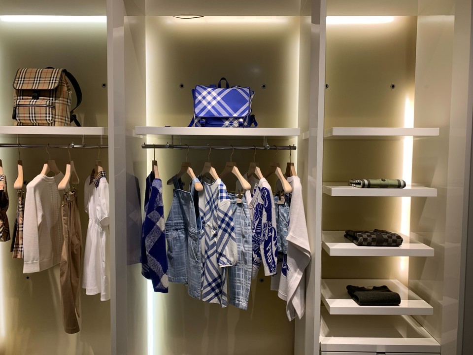 バーバリーチルドレンの店内で、ベージュ色の背景に白い棚に整然と展示された子供服のコレクション。上段にはチェック柄のバックパックが置かれており、ハンガーにはベージュ、白、青のチェック柄のドレスやオーバーオールがかかっている。棚の下段には折りたたまれた黒色の子供服が綺麗に積まれています。