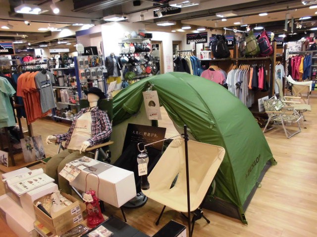 アウトドア用品店の店内ディスプレイで、緑色のキャンプ用テントが設置されている。テントの横にはマネキンがアウトドアウェアを着ており、椅子やランタンなどのキャンプ用品が周囲に展示されている。背景には衣類やバックパックが陳列されている棚が見える。