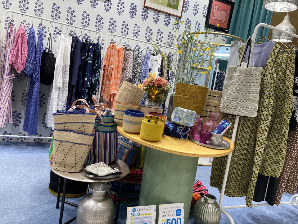 色鮮やかな服やバッグが展示されているテーブルの周りには、さまざまな模様のドレスやチュニックが掛けられています。