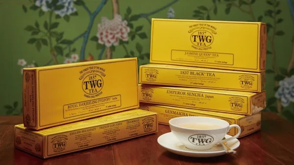 木製のテーブルの上に、黄色いパッケージのTWGティーが積み重なっており、手前にはTWGのロゴ入りティーカップが置かれている。背景には花柄の壁紙が飾られ、高級感のあるティータイムを演出している画像。