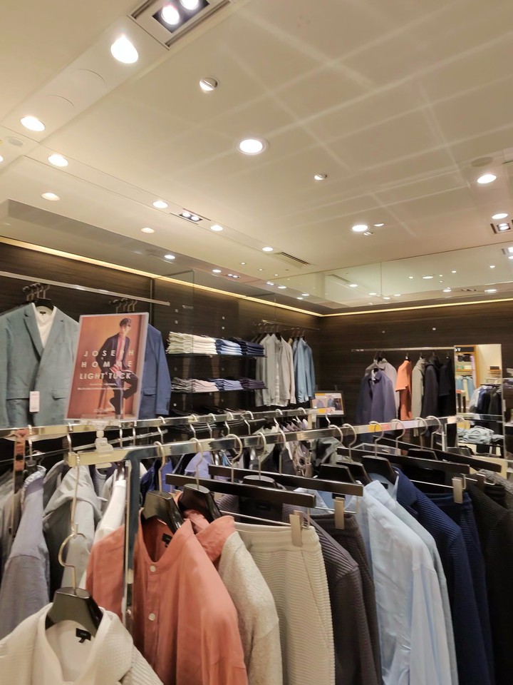 上品なデザインのJOSEPH HOMMEの店内で、洗練されたカジュアルウェアが充実している。前景には色とりどりのシャツが、背景にはポスターと棚に並べられたシャツやジャケットがある。