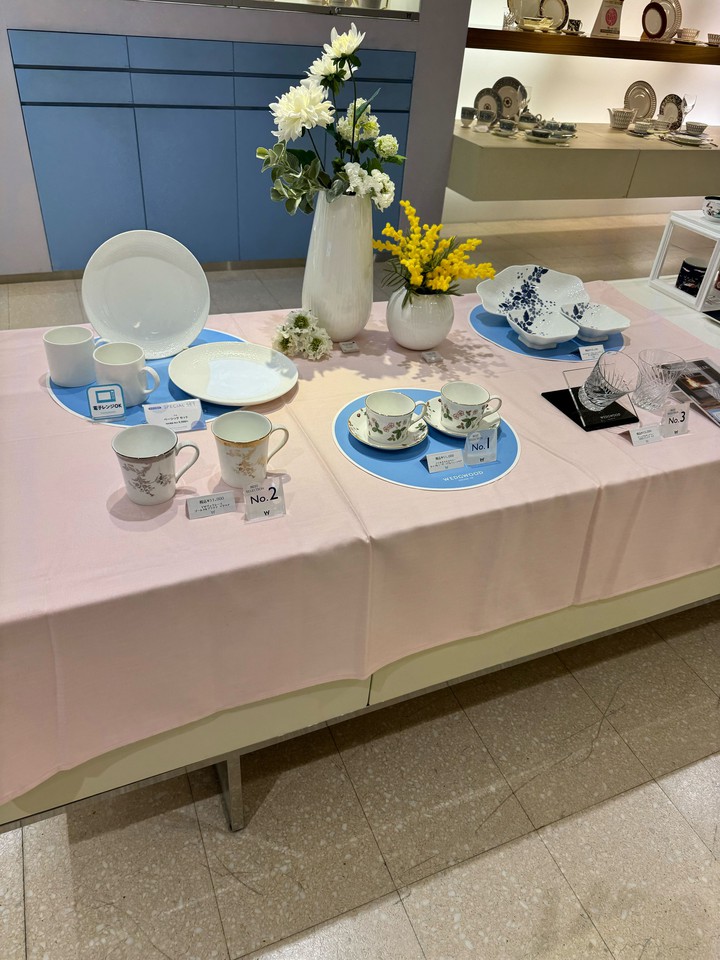 ウェッジウッド ピンクのテーブルクロスの上に展示されたウェッジウッドの食器類。白い花瓶にアレンジされた白と黄色の花が中心に飾られ、その周りには繊細なパターンのティーカップセットや皿が並んでいる。背景には他の食器セットが見える。