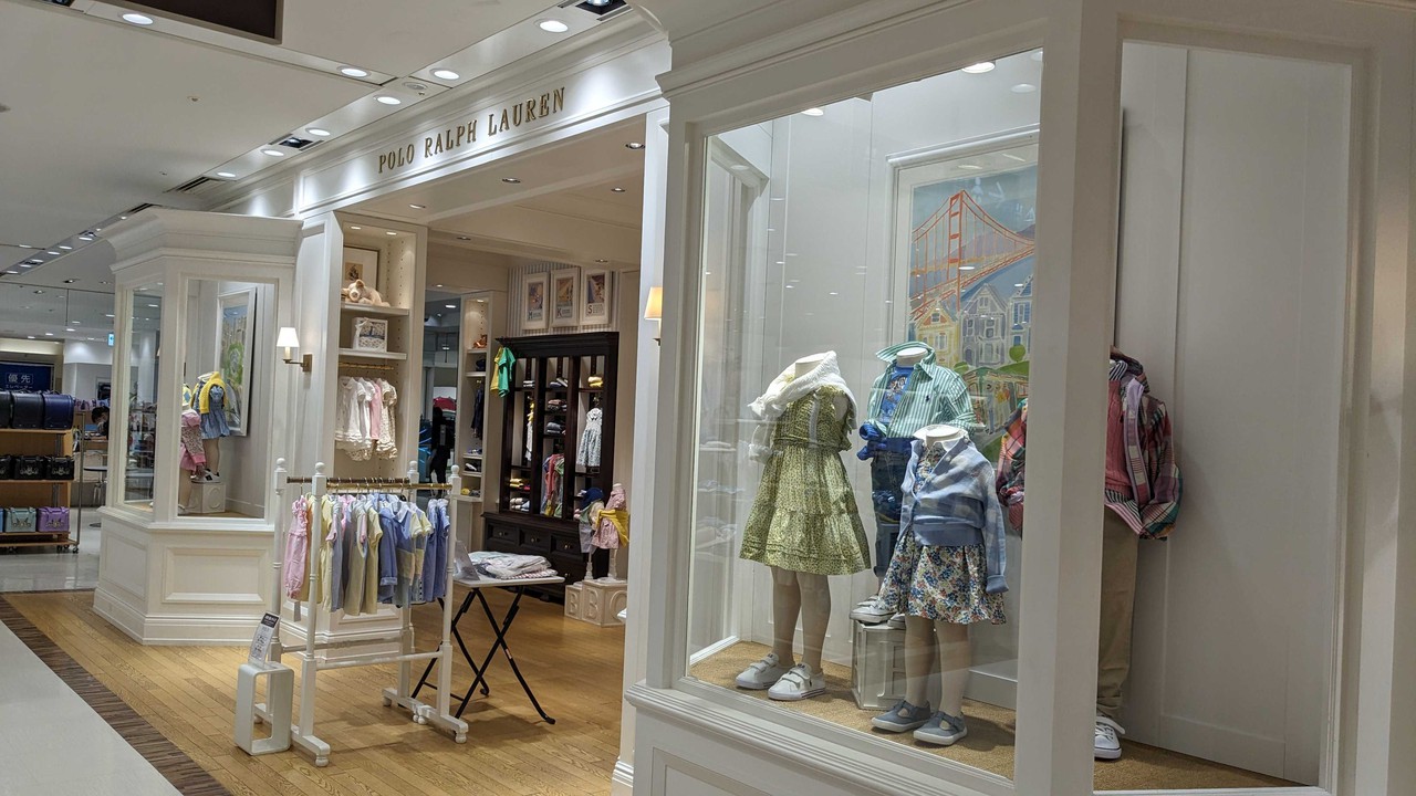 ポロラルフローレンチルドレンの店舗入り口とショーウィンドウ。エレガントな白とベージュの内装で、明るい照明の下には春のコレクションの子供服が展示されています。