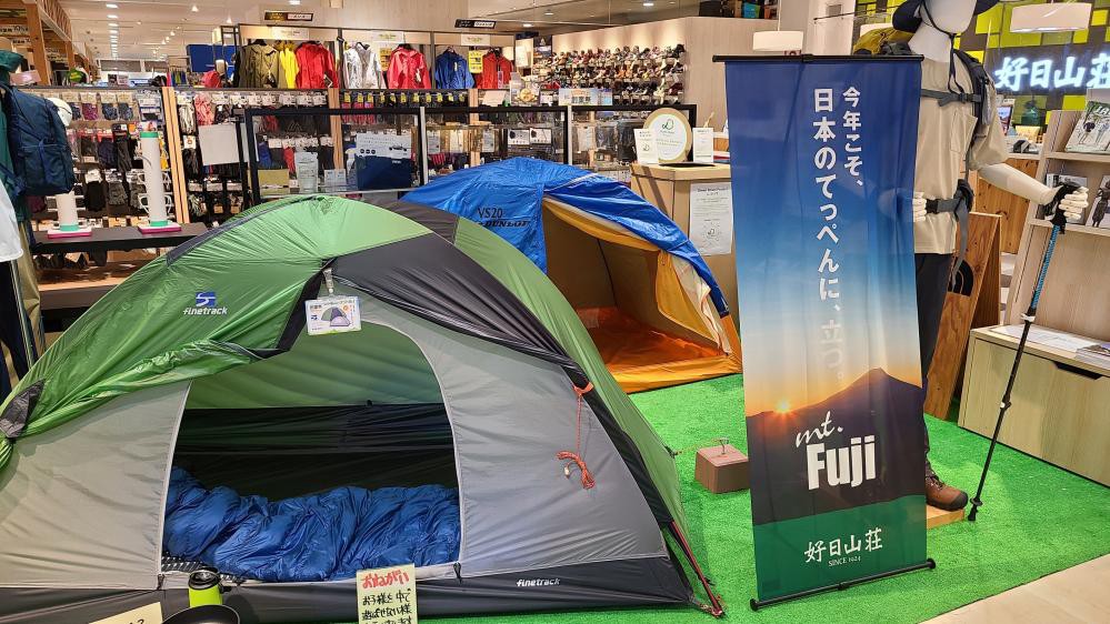 アウトドアショップの展示エリアの画像。緑色のテントと青いテントが並べて設置されており、それぞれの中には寝袋があり、右側には登山装備を身につけたマネキンが展示されている。背景にはその他のアウトドア用品やウェアが並んでいる。