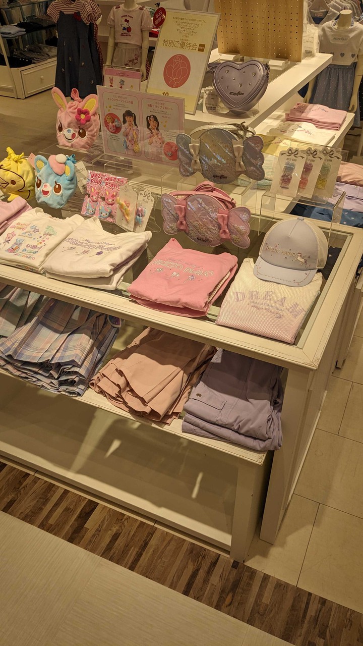 子供服売り場で、ピンクとラベンダーカラーの衣服や小物がディスプレイされています。前面には可愛らしいデザインのキッズ用Tシャツ、キャップ、バッグがあり、背景にはマネキンがドレスやシャツを着て展示されています。売り場の情報を示すポスターも見えます。