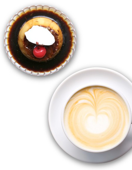 カラメルソースがたっぷりとかかったプリンがあり、その上にはホイップクリームとチェリーがトッピングされている。ハート形のラテアートが描かれたカフェラテが白いカップに入っている。白い背景に、シンプルで美味しそうなデザートとドリンクが強調された画像。