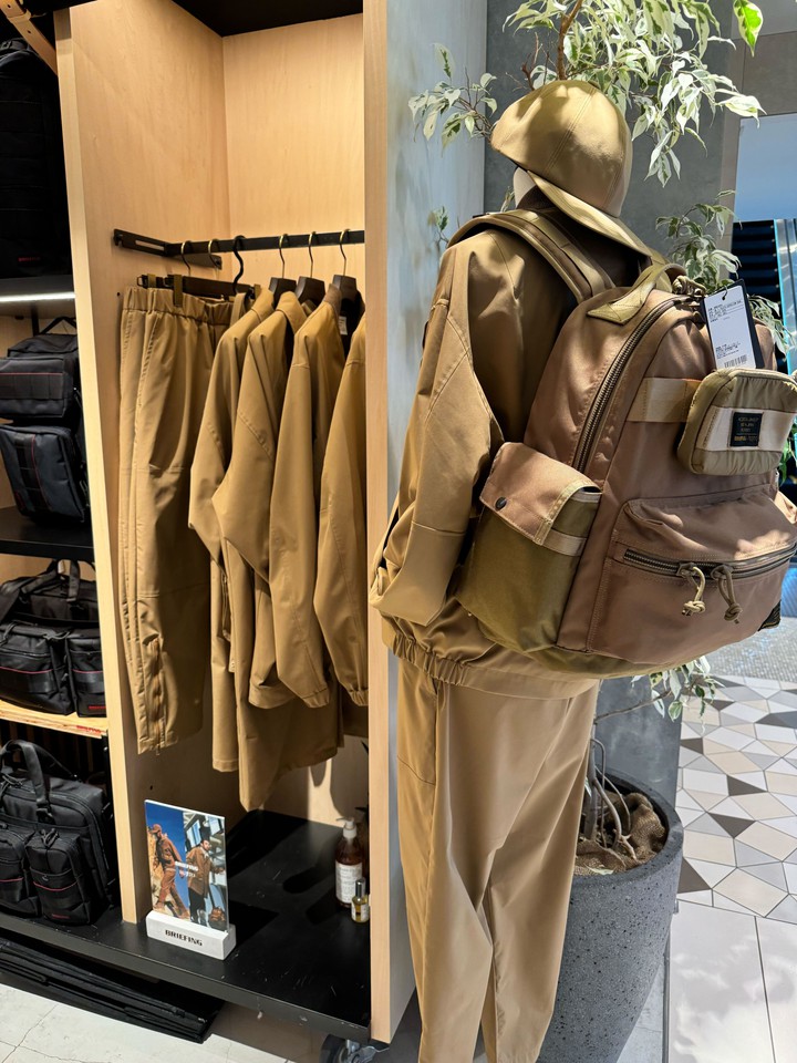 「BRIEFING」の統一されたベージュカラーのアウトフィットとバックパックが展示されている、モダンなファッションコーナー。