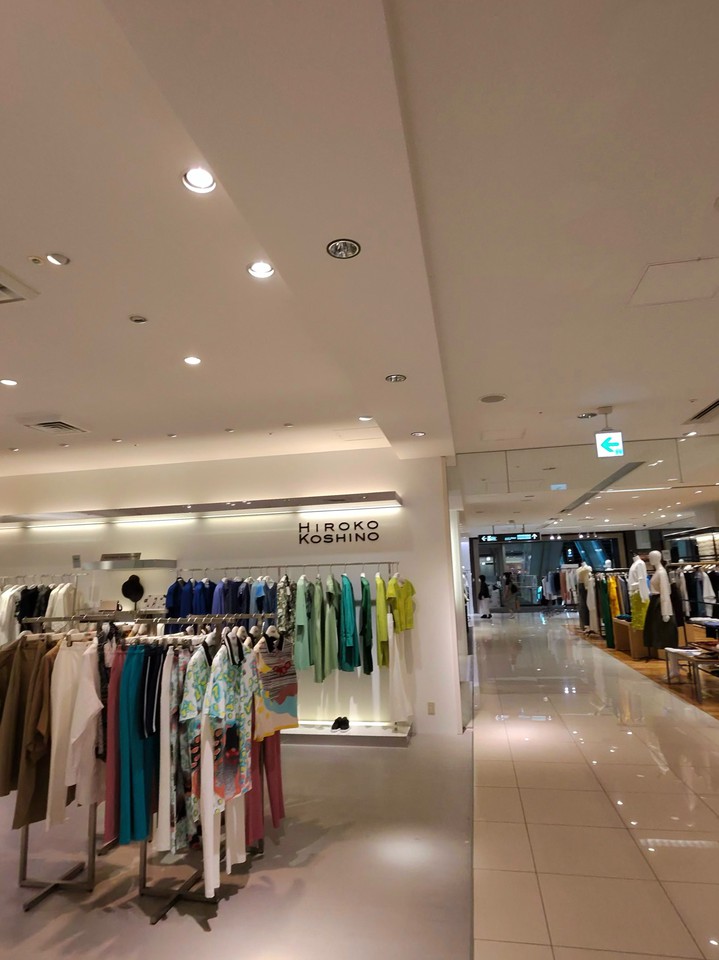 ヒロコ コシノのブティック内部、洗練された明るい空間にカラフルな衣服が並び、前方には緑色のロゴが照明されたブランド名が見える。通路はきらびやかな床が特徴で、左側には目を引く色彩のトップスとボトムスが展示されている。