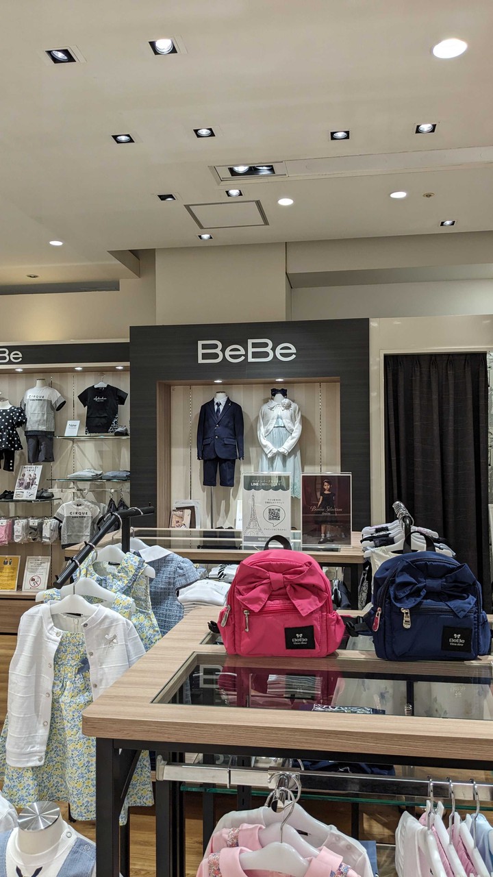 BeBe店内には正統派のネイビーブレザーとフォーマルドレスを着用したマネキンが展示されています。前面のガラスのテーブルの上には、明るいピンクとネイビーのリュックサックが置かれ、子供服の上品で落ち着いたセレクションが見て取れます。