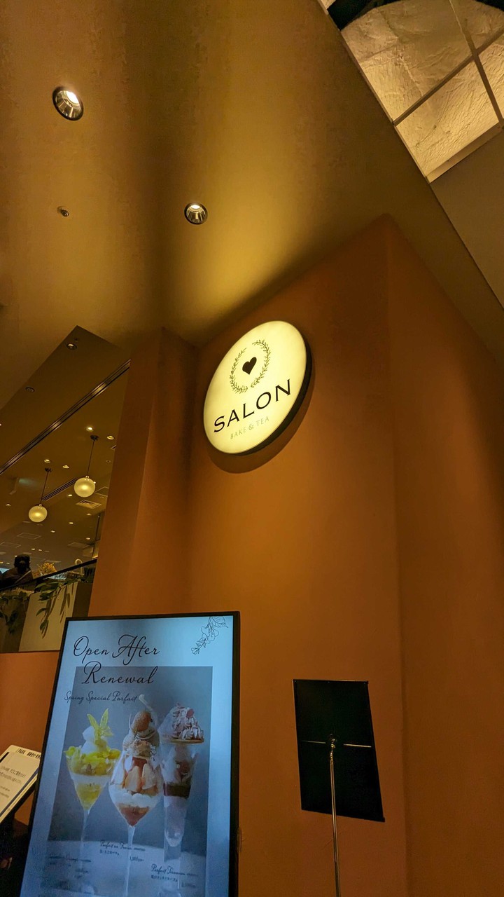 暖色の照明の下で、壁に「SALON BAKE & TEA」と書かれた照明看板があり、その下にはパフェの写真が載ったスタンド看板が見えます。