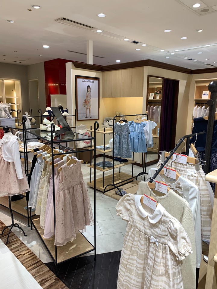 タルティーヌエショコラの店舗内部。ベージュとピンクの女の子用ドレスが前面に展示されている。バーバリーの赤い壁面と、子供用のファッション広告が背景にあり、高級感のある子供服ブティックの雰囲気を演出しています。