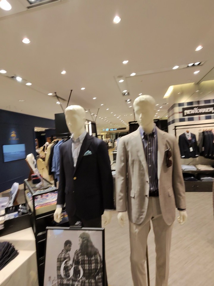 ２体のマネキンがモダンなメンズスーツを着用している店内。左側のマネキンはダークネイビーのスーツに水色のチェック柄シャツ、右側のマネキンはベージュのスーツにストライプのシャツを合わせている。