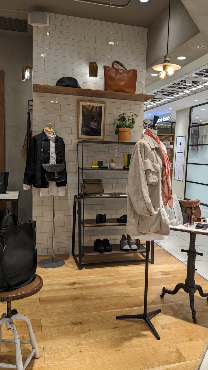 カジュアルな服装がマネキンに展示されているスロウ オーセンティック グッズ ストアの店内の一角。背景にはレザーバッグが棚に掛けられており、天井の照明が商品を明るく照らしている。
