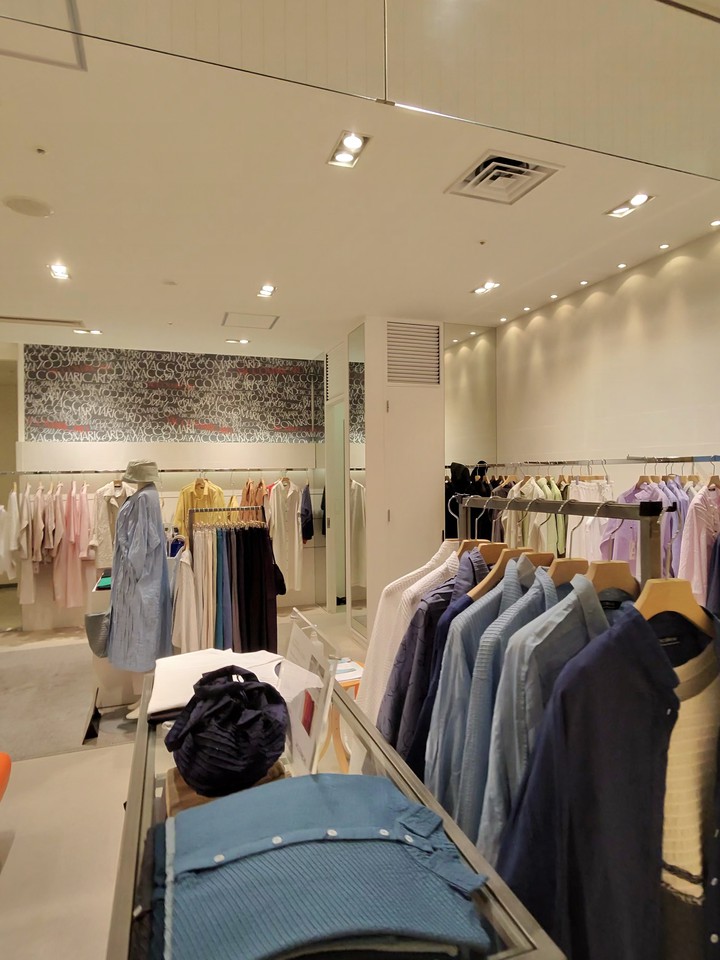 明るくモダンな雰囲気のブティック内部、前面のディスプレイにはさまざまなブルーシャツが置かれ、奥のラックにはカラーパレットのパステルトーンの服が陳列されている。壁には「YACCOMARICARD」の文字が繰り返された装飾的なアートワークがある。